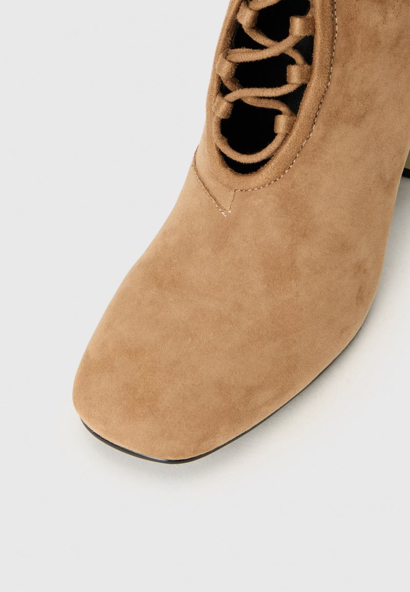 Beige Wildleder-Ankle-Boot mit eckiger Zehenpartie, ausgestattet mit einem geschnürten Cutout-Design und einer glatten Textur, das einen eleganten Absatz präsentiert.