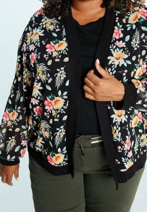 Femme portant un cardigan noir à motifs floraux sur un haut noir et un pantalon vert olive, une main tenant le cardigan ouvert.