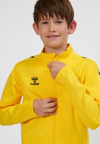 Gul zip-up jacka i slät tyg med hög krage, med svart logotyp och detaljer på axlarna.