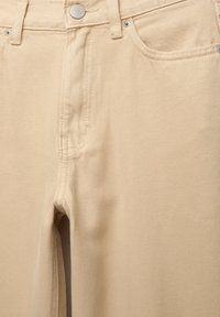 Jean denim beige vue de face montrant un bouton métallique, des passants de ceinture et des poches avant avec des détails de couture visibles.