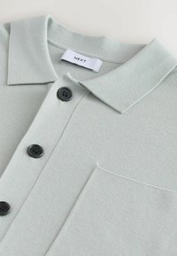 Svijetlozeleni pleteni majica s ravnim ovratnikom, tri crne gumbe i džepom na prsima. Tkanina ima rebrastu teksturu. Brand etiketa: NEXT.