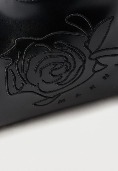 Μαύρη δερμάτινη επιφάνεια με ανάγλυφο περίγραμμα ενός τριαντάφυλλου και μερικώς ορατό κείμενο "MARC" κάτω από το σχέδιο.