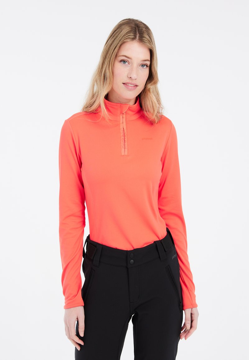 Camicia a maniche lunghe e colletto alto di un vivace arancione, con zip frontale a metà. Materiale morbido ed elastico, indossata con pantaloni neri e un logo minimalista.