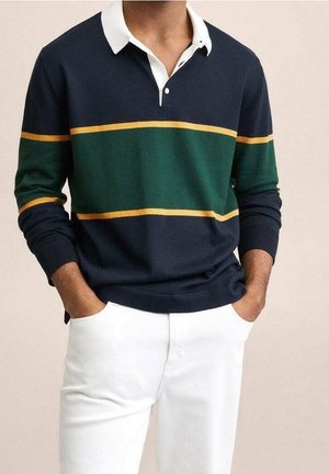 Mężczyzna ubrany w granatowo-zielono-żółtą koszulkę polo z długim rękawem w paski, z białym kołnierzykiem i białe spodnie, z rękami w kieszeniach.