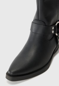 Bota de tobillo de cuero negro con punta afilada, detalle de correa simple y acentos en metal plateado, con suela de goma plana.