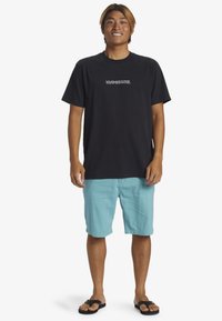 Svart bomullströja med vit grafisk text, parat med ljusblå shorts och svarta flip-flop. Lättviktig, avslappnad sommaroutfit.