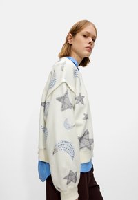 Felpa bianca con motivi di stelle e lune, vestibilità oversize, polsini a coste, indossata sopra una maglietta blu. Tessuto morbido, design casual.