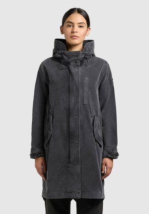 Ung kvinde med mørkt hår iført en lang, mørkegrå hættetrøje med knapper på ærmekanterne og forlommer, stående mod en ensfarvet baggrund.