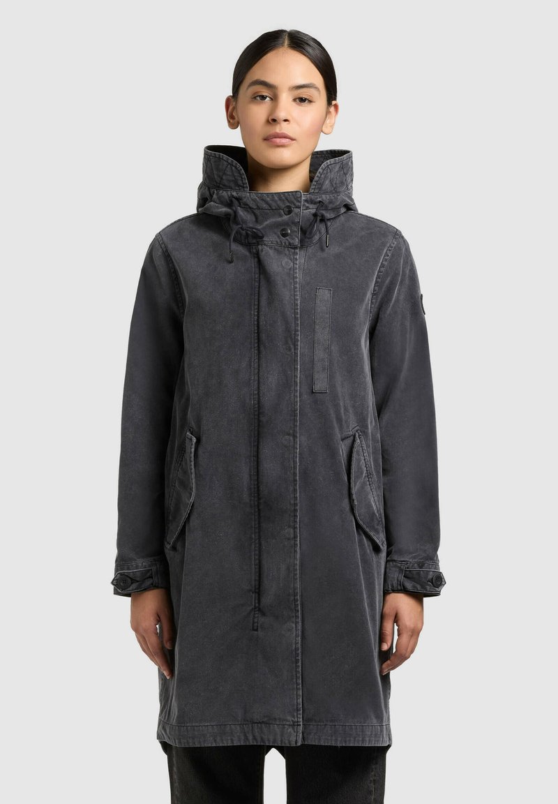 Giovane donna con capelli scuri che indossa un lungo cappotto con cappuccio grigio scuro, polsini con bottoni e tasche frontali, in piedi davanti a uno sfondo semplice.