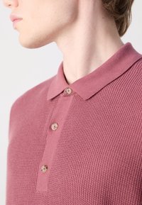Personne portant un polo texturé rose poudré à trois boutons, vue du cou au haut du torse, tournée vers la gauche sur un fond uni.
