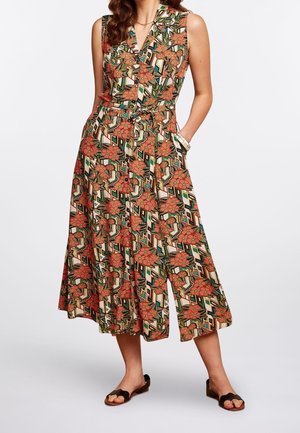 Woman wearing sleeveless midi dress with floral and geometric pattern, standing with hands in pockets, wearing black flat sandals.