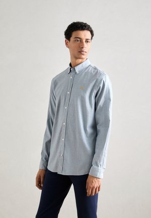 Camisa azul clara de botones con un cuello corto, que presenta un logo dorado bordado en el pecho y mangas largas, hecha de una tela suave.