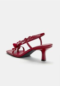 Sandales en cuir verni rouge avec un talon fin, un bout carré et un design de brides entrecroisées avec un accent floral. Texture lisse sur l'ensemble.