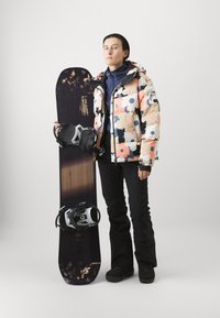 Fodrad, flerfärgad blommig jacka över en blå topp, svarta snöbyxor och stövlar, håller en mörk snowboard med bindningar, mot en neutral bakgrund.