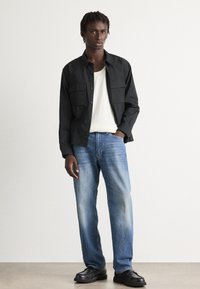 Svart skjorte med knapper og to brystlommer, hvit ribbestrikket skjorte under, lyseblå denimjeans og svarte sko, mot en nøytral bakgrunn.