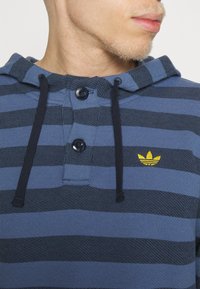 Blå och marinrandig hoodie med en krage med knappar, mörka snoddar och en liten gul broderad logotyp på bröstet.