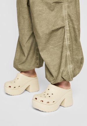 Füße, die beige klobige Plateau-Clogs tragen, kombiniert mit lockeren, hellbraunen, weiten Hosen vor grauem Hintergrund.