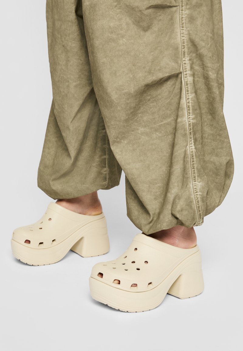 Pieds portant des sabots beiges à plateforme épaisse associés à un pantalon ample marron clair sur un fond gris.