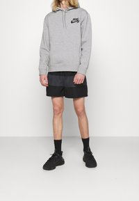 Sudadera con capucha gris y logo negro, combinada con pantalones cortos a rayas en negro y gris. Zapatillas negras y calcetines oscuros completan el conjunto.