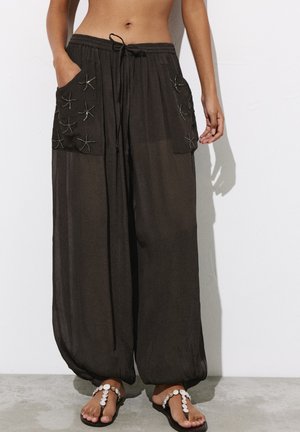 Pantalon noir ample avec de grandes poches avant décorées d'étoiles de mer cousues, porté avec des sandales ornées de coquillages et un ventre découvert.