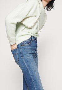 Suéter esponjoso verde claro, jeans azul oscuro con bolsillo lateral, y un bolso blanco con un diseño curvado y detalle de cordón.