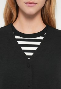 Cardigan noir avec une finition texturée, doté d'un col en V et de deux boutons. En dessous, un haut blanc à rayures noires avec un col rond.
