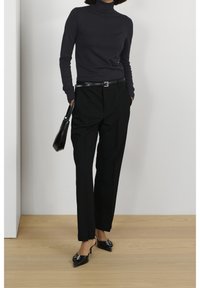 Czarny sweter z golfem, dopasowane czarne spodnie z paskiem oraz czarne, szpiczaste mules na obcasie. Minimalistyczna torebka jako dodatek. Neutralne tło.