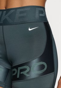 Szorty Nike Pro w ciemnozielonym kolorze. Posiadają szeroki pas z jasnoszarym napisem oraz małym białym logo z przodu. Gładka faktura.