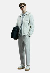 Veste blanche légère avec capuche, poches zippées ; chemise grise en dessous ; pantalon gris clair ; lunettes de soleil noires ; baskets blanches avec accents.