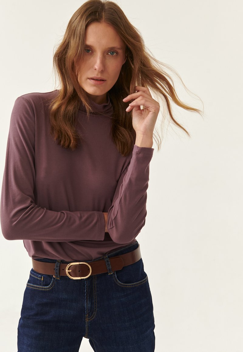 Cămașă cu mânecă lungă, guler înalt, în nuanță de mauve, având o textură fină. Este asortată cu blugi de culoare albastru închis și o curea maro cu o buckle de aur.
