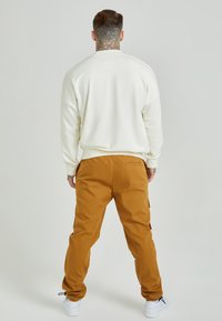 Krämfärgad sweatshirt med ribbade manschetter och avslappnad passform, i kombination med bruna cargobyxor med sidofickor och en lös silhuett. Vita sneakers.