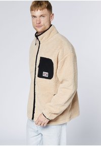 Junger Mann trägt eine beige Fleecejacke mit einer schwarzen Reißverschlusstasche auf der Brust und hellblaue Jeans, steht vor einem einfarbigen Hintergrund.