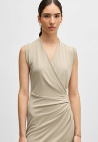 Beige sleeveless dress with a wrap design, featuring gathers on the side and a smooth texture. Simple, minimalist silhouette.