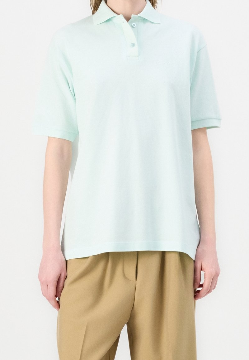 Personne portant un polo manches courtes vert menthe clair et un pantalon beige ample, debout devant un fond blanc uni.