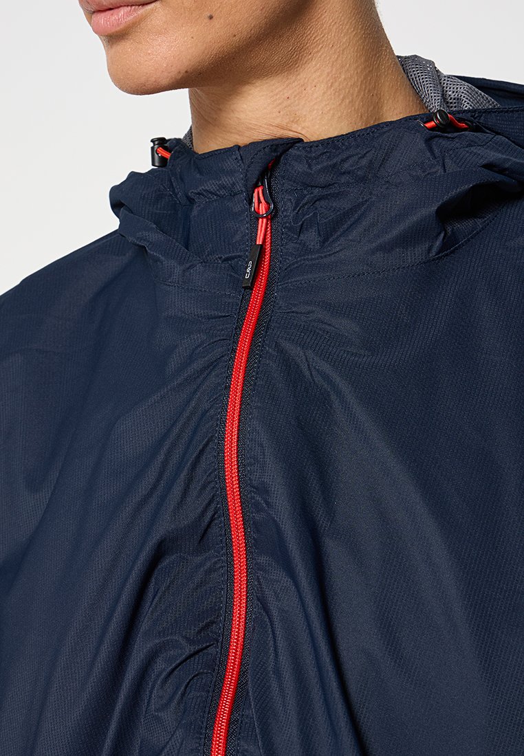 Primo piano di una persona che indossa una giacca a vento blu navy con zip rossa e lacci regolabili al colletto.