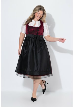Mujer sonriente con un vestido tradicional burdeos y negro con mangas de encaje y tacones negros, posando con un pie levantado.