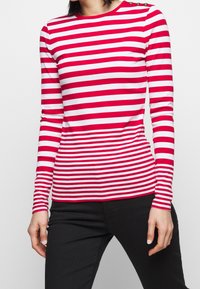 Czerwona i biała w paski koszula z długim rękawem, okrągłym dekoltem i guzikami na ramieniu, wykonana z gładkiego, elastycznego materiału.