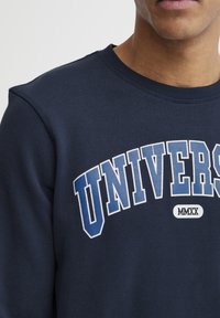 Marinblå sweatshirt med ribbad hals, med vit och ljusblå text "UNIVERS" och en liten "MMXX" detalj i mitten. Mjuk tygkänsla.