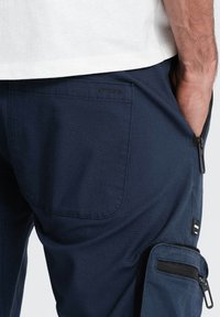 Pantalons cargo bleu marine avec une poche arrière, une poche zippée sur le côté et un tissu texturé. Comprend un logo près de la taille et des accents zippés.