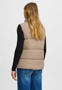 Beige vadderad väst med hög krage, med kviltade sektioner. Matchad med en svart långärmad tröja och löst sittande blå jeans.