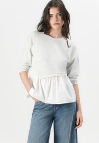 Sudadera gris claro con un dobladillo peplum blanco, que presenta mangas largas y un cuello redondo. La tela es suave y tiene un corte relajado.