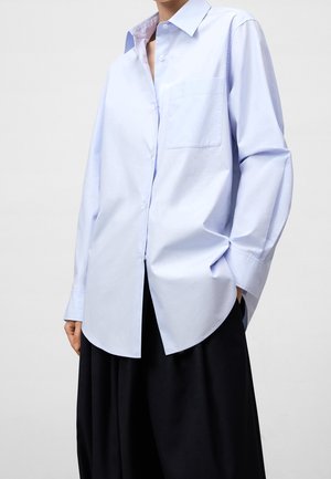 Personne portant une chemise ample bleu clair à boutons avec poche poitrine et un pantalon noir à jambes larges, main dans la poche, sur fond uni.
