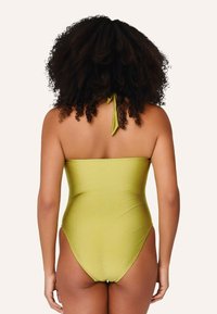 Olivovo zelené jednodielne plavky s viazaním za krkom, hladká textúra, vysoké rozparky na nohách a minimálne zakrytie chrbta. Žiadne výrazné vzory ani kovové súčasti.