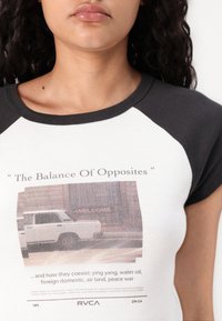 Camiseta con cuerpo blanco y mangas raglán negras. Presenta una impresión gráfica de un coche y el texto: "El Equilibrio de los Opuestos" en colores apagados.