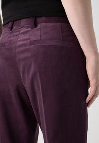 Pantalons en velours côtelé violet avec poches arrière, présentant un motif texturé et une coupe ajustée. Le tissu a une surface douce et côtelée.