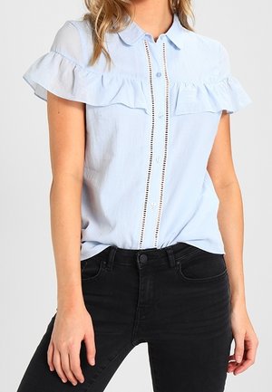 Kvinde iført en lyseblå kortærmet bluse med flæsedetalje og sorte jeans med høj talje, stående med hænderne afslappet ned langs siderne.