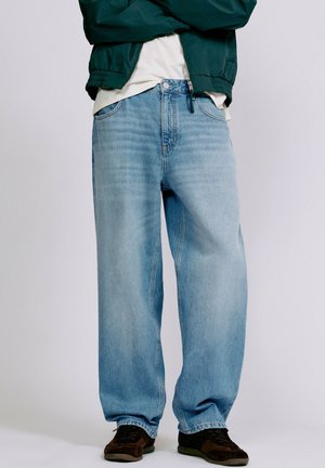 Personne portant un jean bleu clair ample, des baskets noires, une chemise blanche et une veste à zip verte, bras croisés.