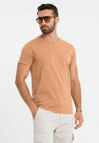 T-shirt in cotone beige con collo rotondo, maniche corte e vestibilità slim, abbinata a occhiali da sole neri oversize in un ambiente soleggiato.