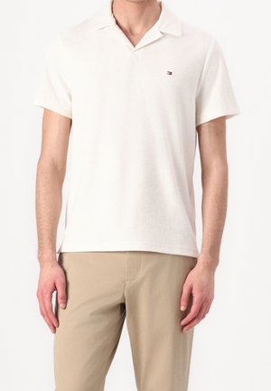 Polo blanc à manches courtes avec col et petit logo brodé sur la poitrine, porté avec un pantalon beige.