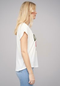 T-shirt en coton blanc avec des manches roulées, affichant un texte graphique vert et rouge sur le devant. Associé à un jean en denim bleu clair.